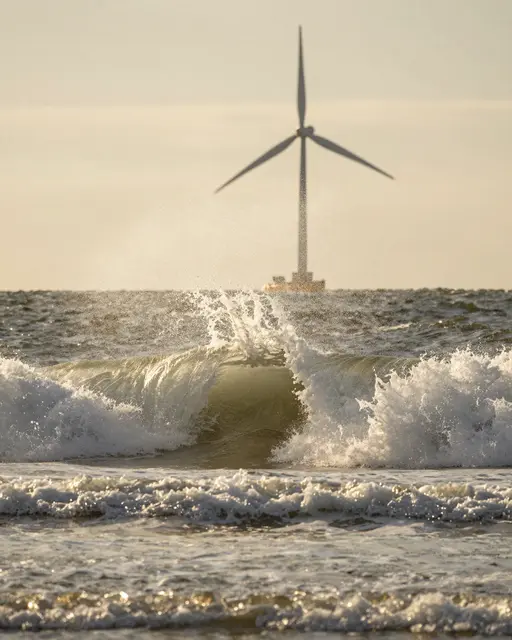 A breaking surf wave catches golden backlight with an offshore wind turbine visible on the horizon