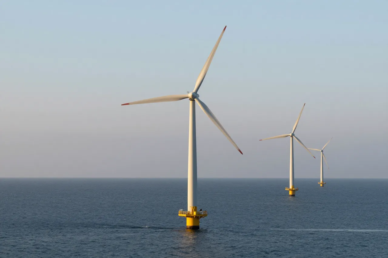 Three offshore wind turbines stand in a line on calm ocean water under a soft hazy sunrise sky