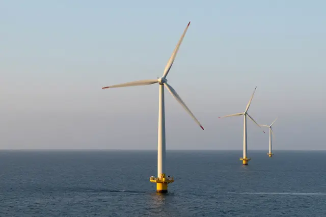 Three offshore wind turbines stand in a line on calm ocean water under a soft hazy sunrise sky