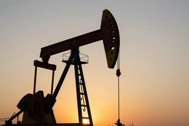 An oil pumpjack silhouette stands against a warm gradient sunset sky with the low sun framed under its support leg