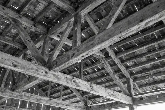 Looking up at crisscrossed weathered wooden barn roof beams with light streaming through plank gaps in black and white