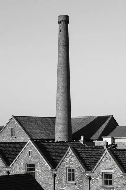 Black and white view of a tall brick factory chimney rising above rows of brick building roofs under an overcast sky