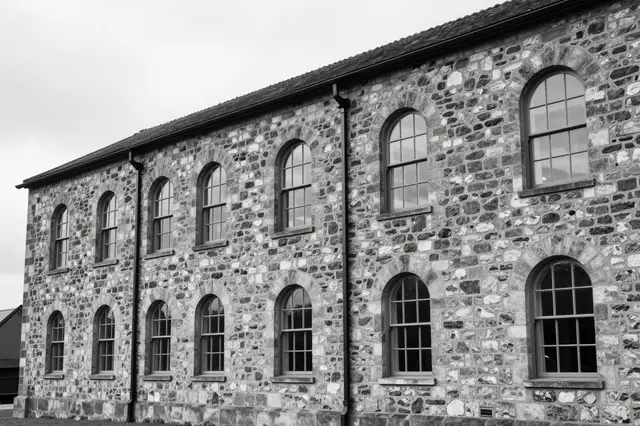 Black and white view of an old rough stone mill building with rows of arched windows under overcast light