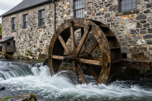 Long exposure view of a wooden waterwheel set in rushing mill race water beside a historic stone mill building