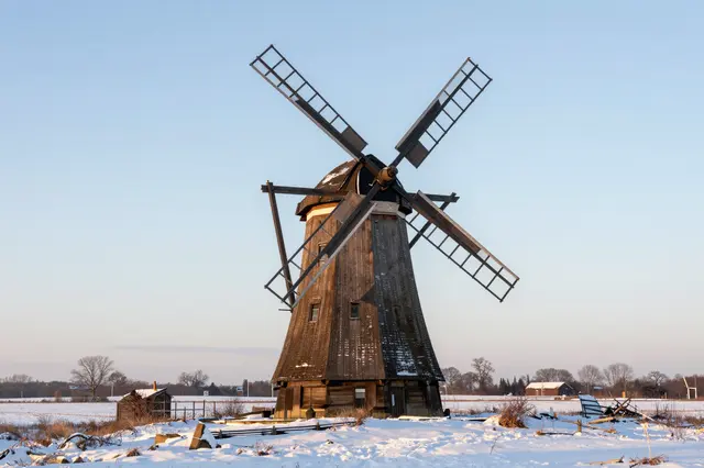 Weathered wooden old windmill stands in a snow covered open field under a pale clear winter sky