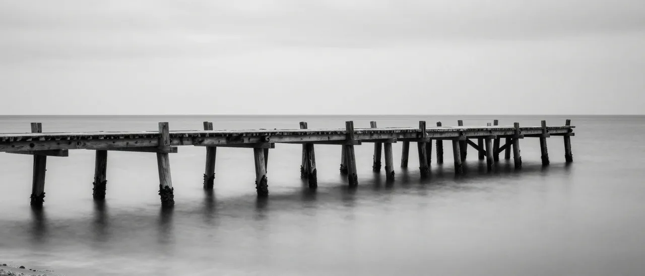 Black and white long exposure view of an old wooden jetty stretching across glassy calm sea under an overcast sky