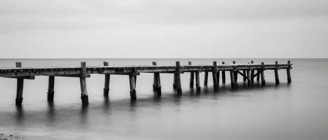 Black and white long exposure view of an old wooden jetty stretching across glassy calm sea under an overcast sky