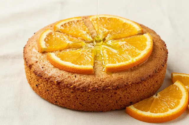 Golden olive oil cake topped with fresh orange slices as oil pours over it, resting on a light linen cloth