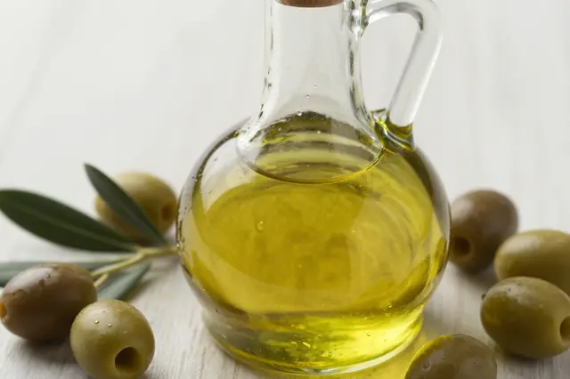 A glass olive oil cruet sits surrounded by fresh olives and an olive sprig on a light wooden surface