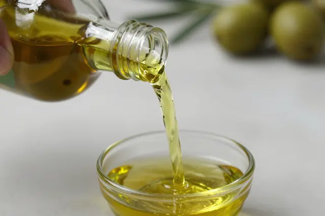 A hand pours golden olive oil from a clear bottle into a small glass dish, with olives blurred in the background.