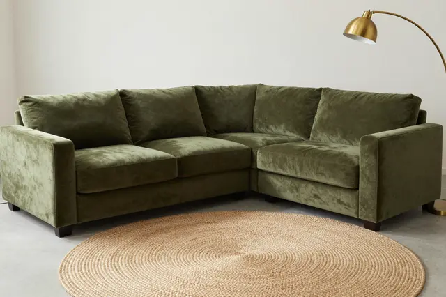 An olive velvet L-shaped sectional sits above a round jute rug next to a brass arc floor lamp in a minimal living room.