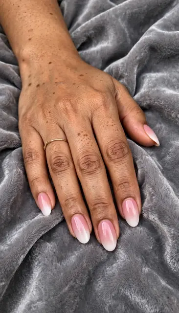 A South Asian woman's hand with pink to white ombre oval nails rests on soft grey velvet.