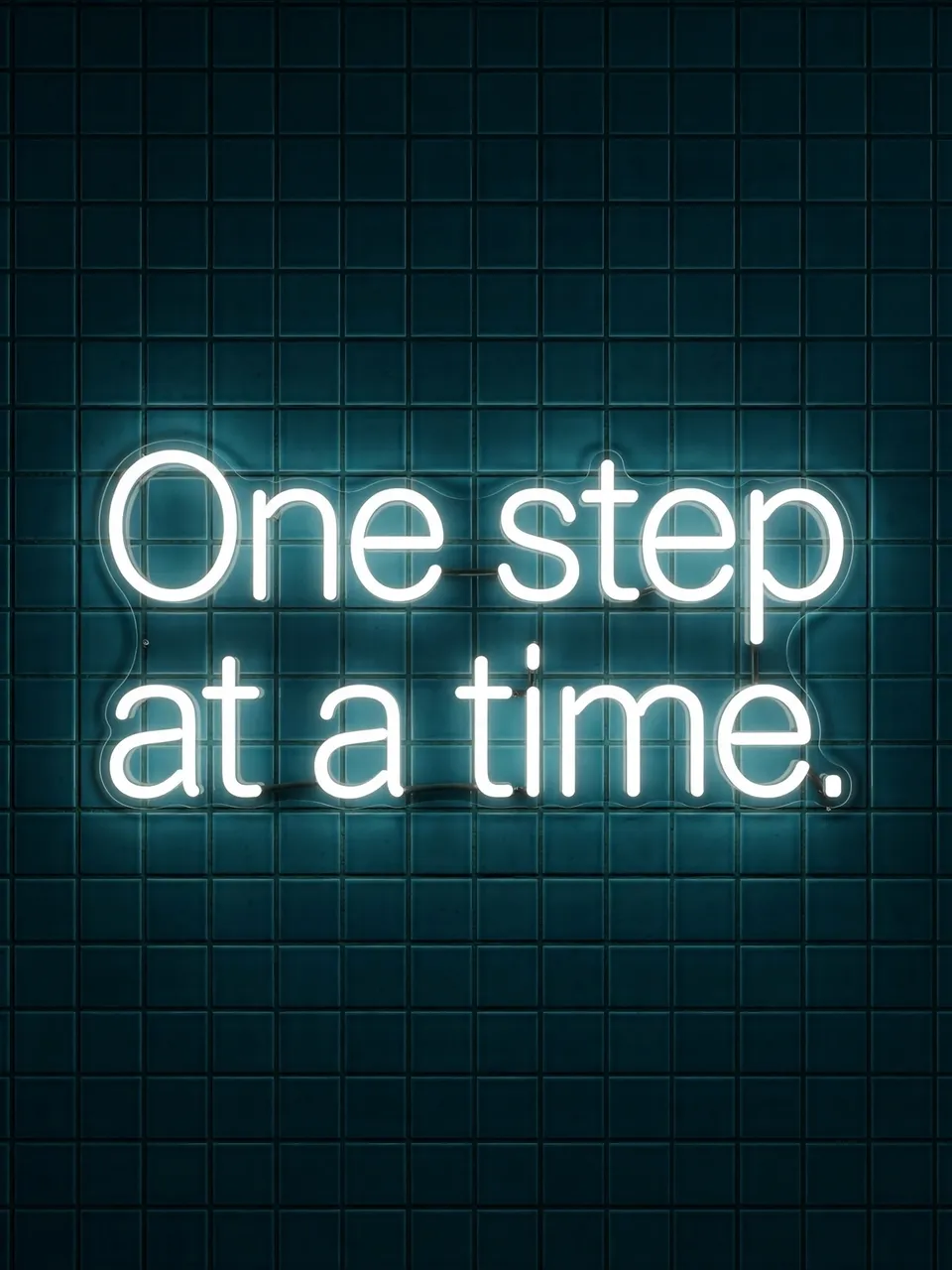 Glowing white neon sign reading "One step at a time." mounted on a dark teal tiled wall