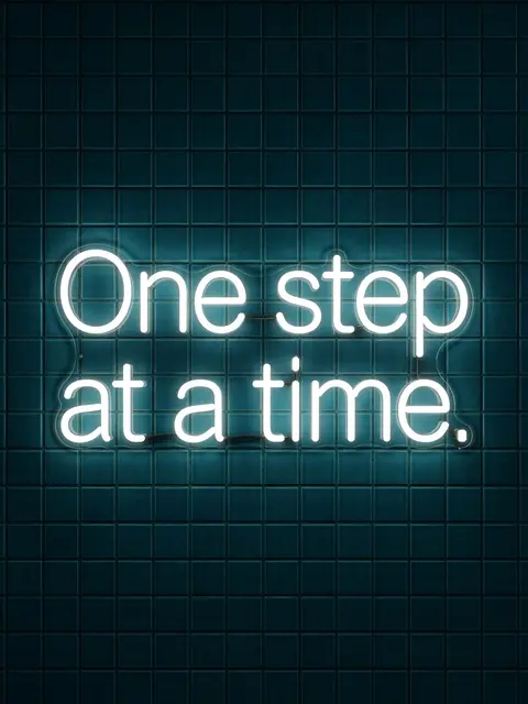 Glowing white neon sign reading "One step at a time." mounted on a dark teal tiled wall