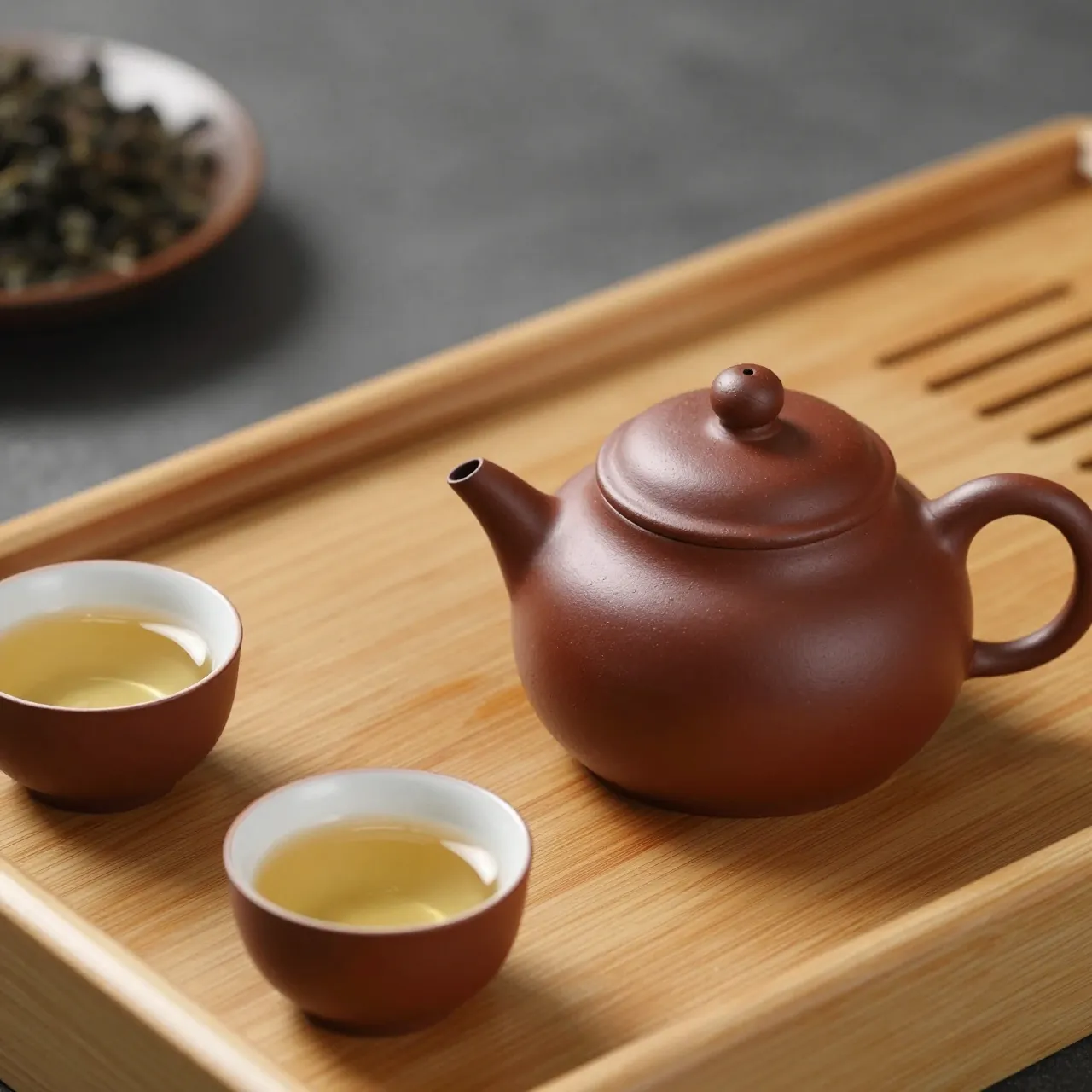 A brown clay teapot with filled tea cups rests on a slotted wooden tea tray next to a bowl of loose oolong tea.