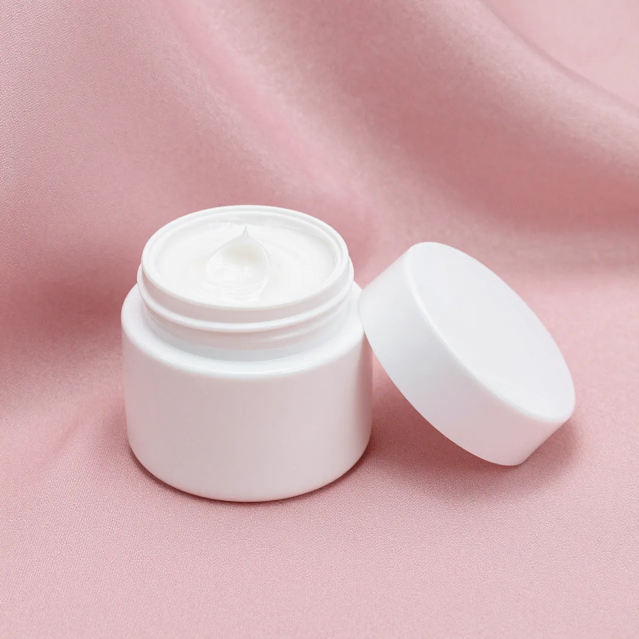 An open white cream jar with its lid resting beside it on soft pink silk fabric