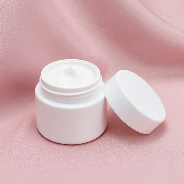An open white cream jar with its lid resting beside it on soft pink silk fabric