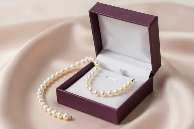 An open purple jewelry gift box holds a pearl necklace resting on soft blush silk fabric.