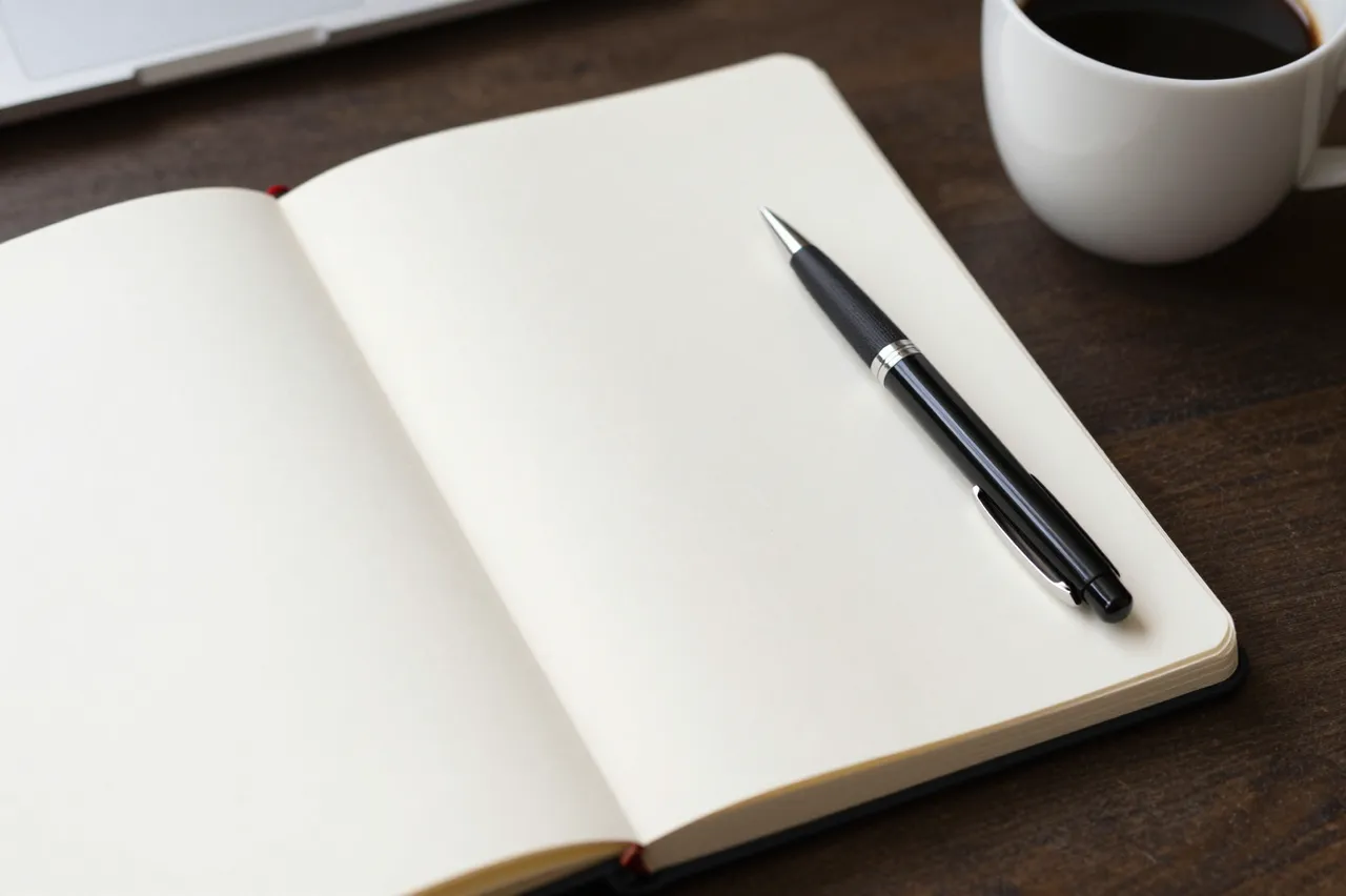 An open blank notebook with a pen sits next to a coffee cup on a dark wood desk.