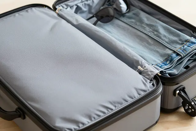 Top view of an open hard-shell suitcase with folded jeans and sunglasses resting on a light wooden surface