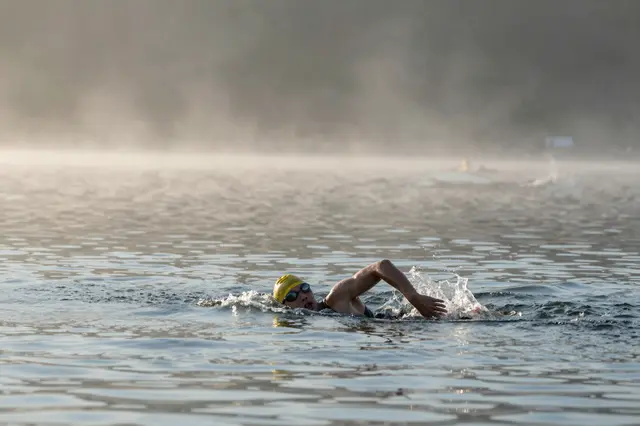 An open-water swimmer with a yellow cap swims across a foggy lake at early dawn.