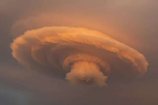 A layered, swirling cloud formation glows warm orange against a muted hazy sky after storm conditions.