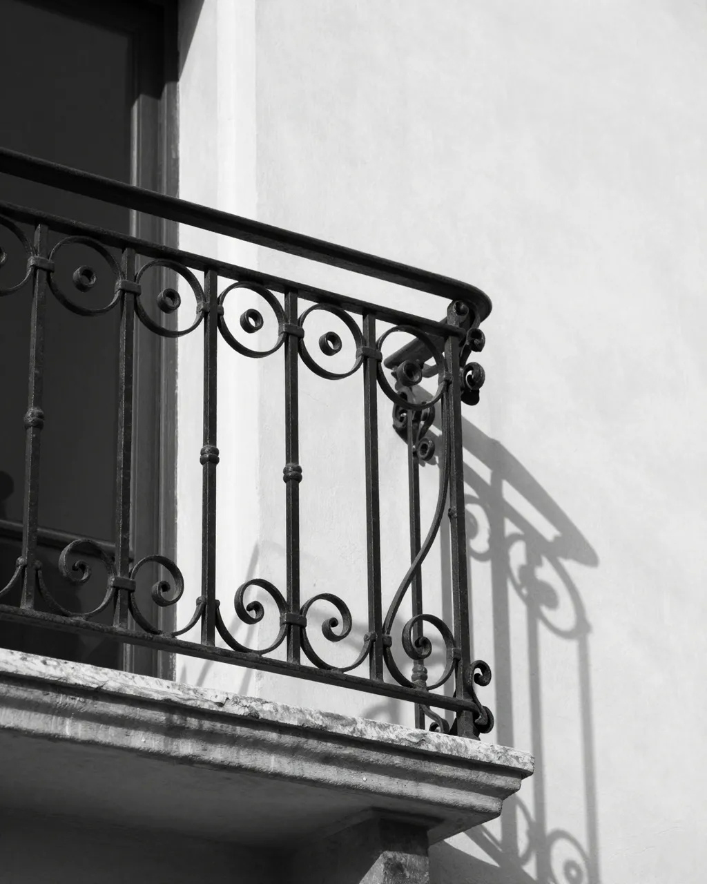 Black and white close-up of an ornate wrought iron balcony railing casting curling shadows on a pale plaster wall