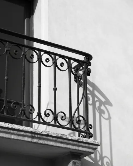 Black and white close-up of an ornate wrought iron balcony railing casting curling shadows on a pale plaster wall