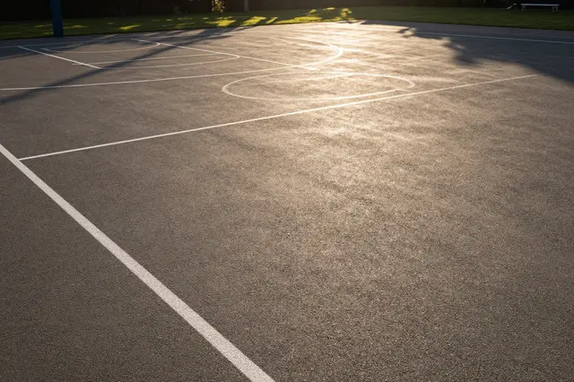 Sunset light casts long tree shadows across an empty paved outdoor basketball court