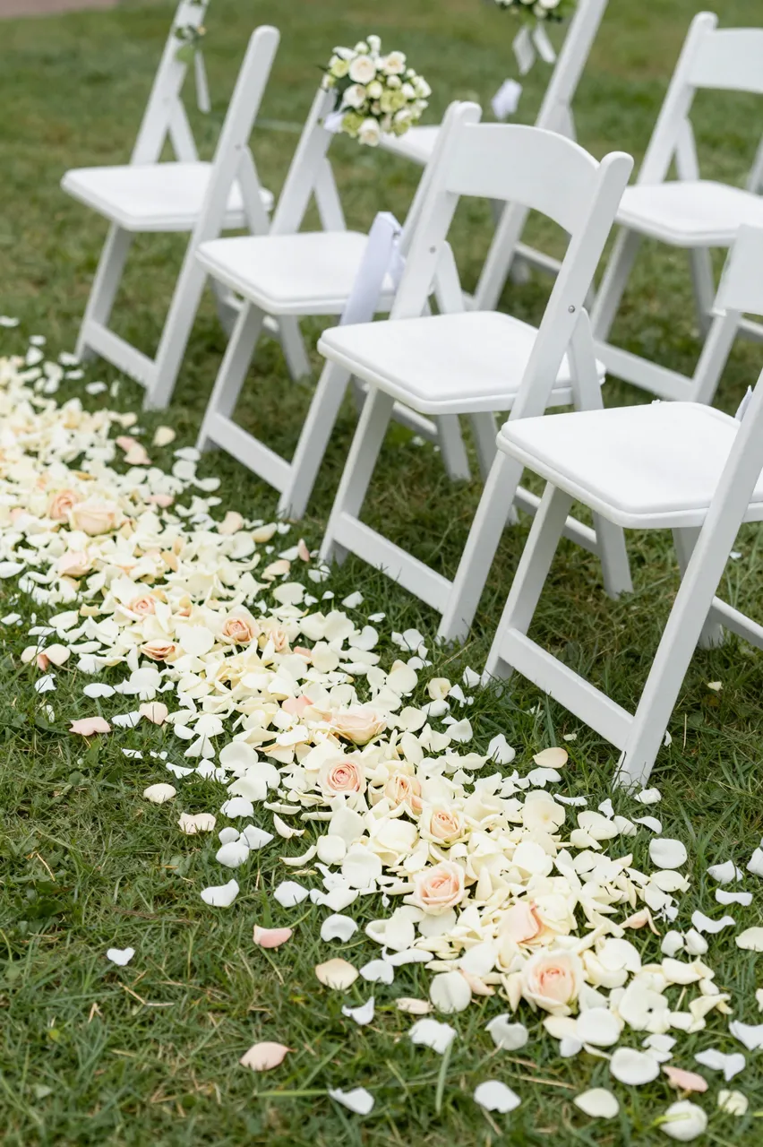 White folding chairs line a grass wedding aisle scattered with white and blush rose petals.