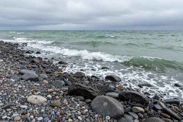 Green cold waves roll onto a rounded dark stone beach under a low gray overcast sky