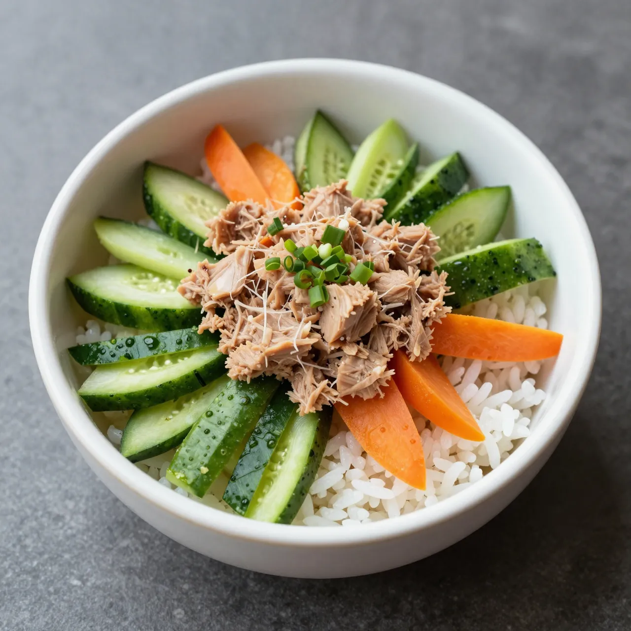 Top down view of a white bowl holding rice, shredded tuna, sliced cucumber, carrot and scallions on a gray countertop