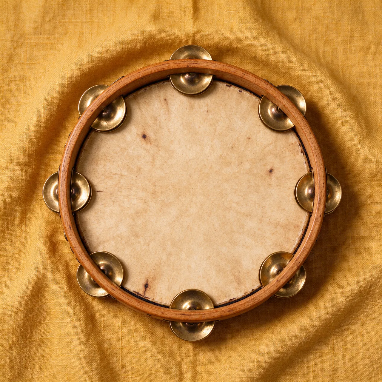 A wooden tambourine with brass jingles rests flat on a textured mustard yellow linen cloth viewed from above.