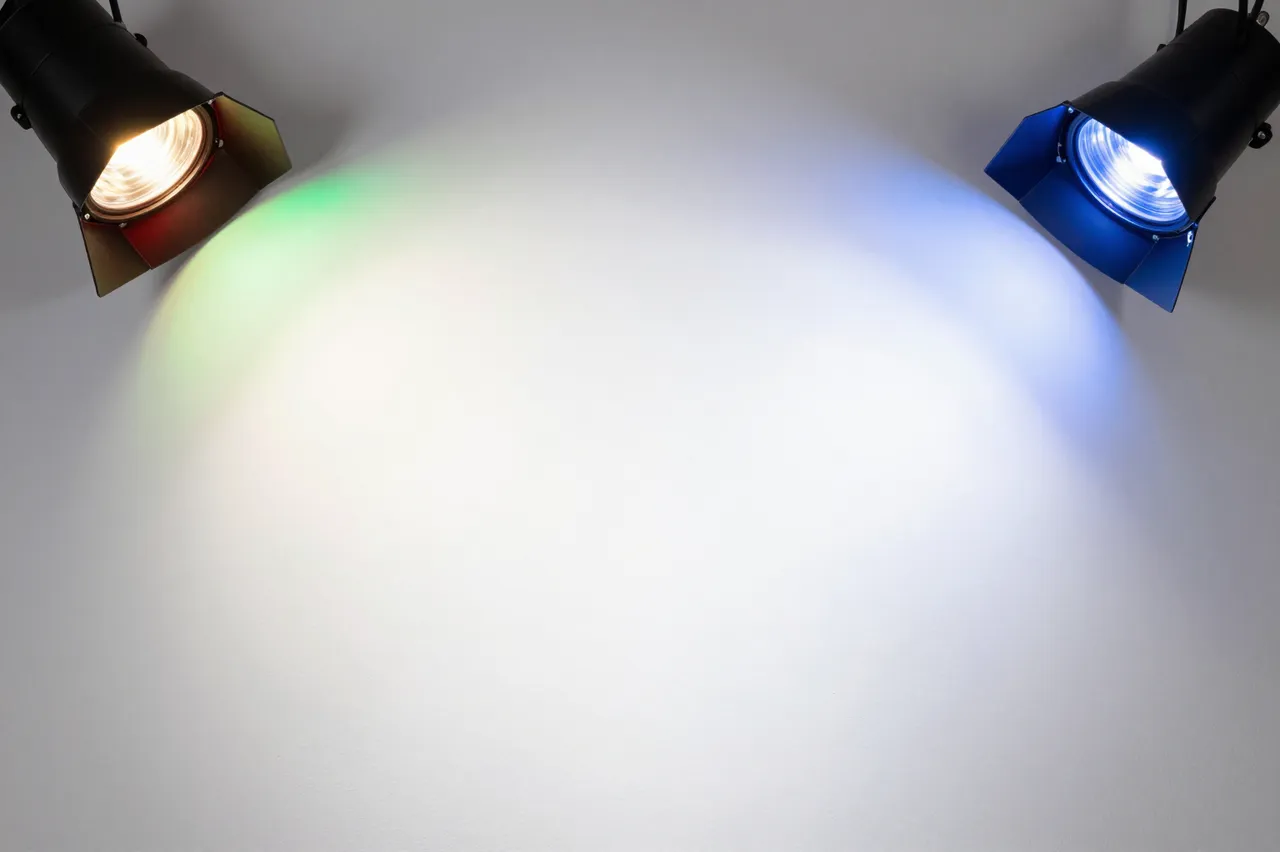 Two stage spotlights cast overlapping red, green and blue light onto a plain white wall with a bright blended center.