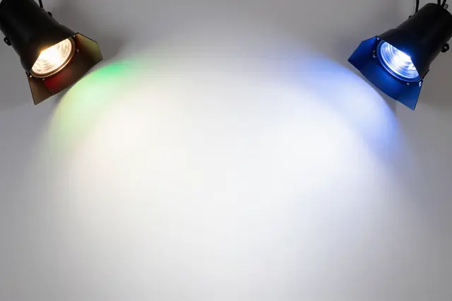 Two stage spotlights cast overlapping red, green and blue light onto a plain white wall with a bright blended center.