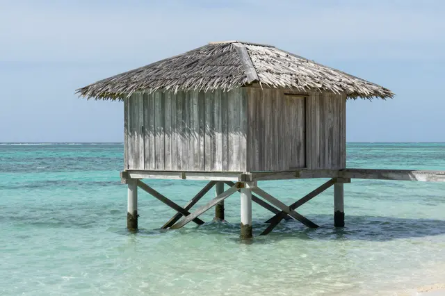 A weathered wooden stilt hut with a thatched roof sits above clear turquoise tropical ocean water.