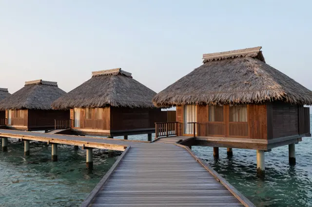 Thatched roof overwater villas connected by a wooden walkway over calm turquoise ocean at dusk