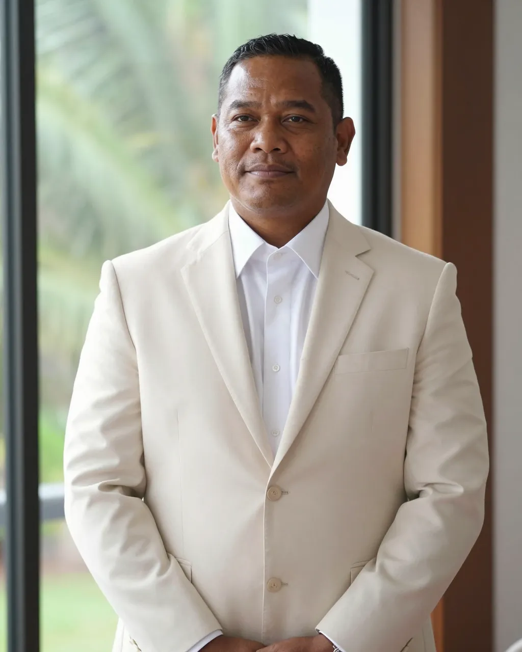 Calm Pacific Islander man in a cream suit stands by a bright window with a soft smile