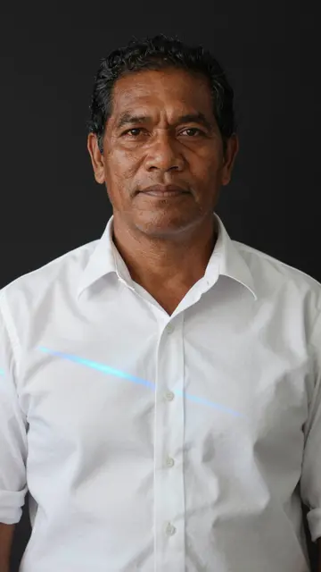 A middle aged Pacific Islander man in a white shirt with a blue projected light line against a dark background
