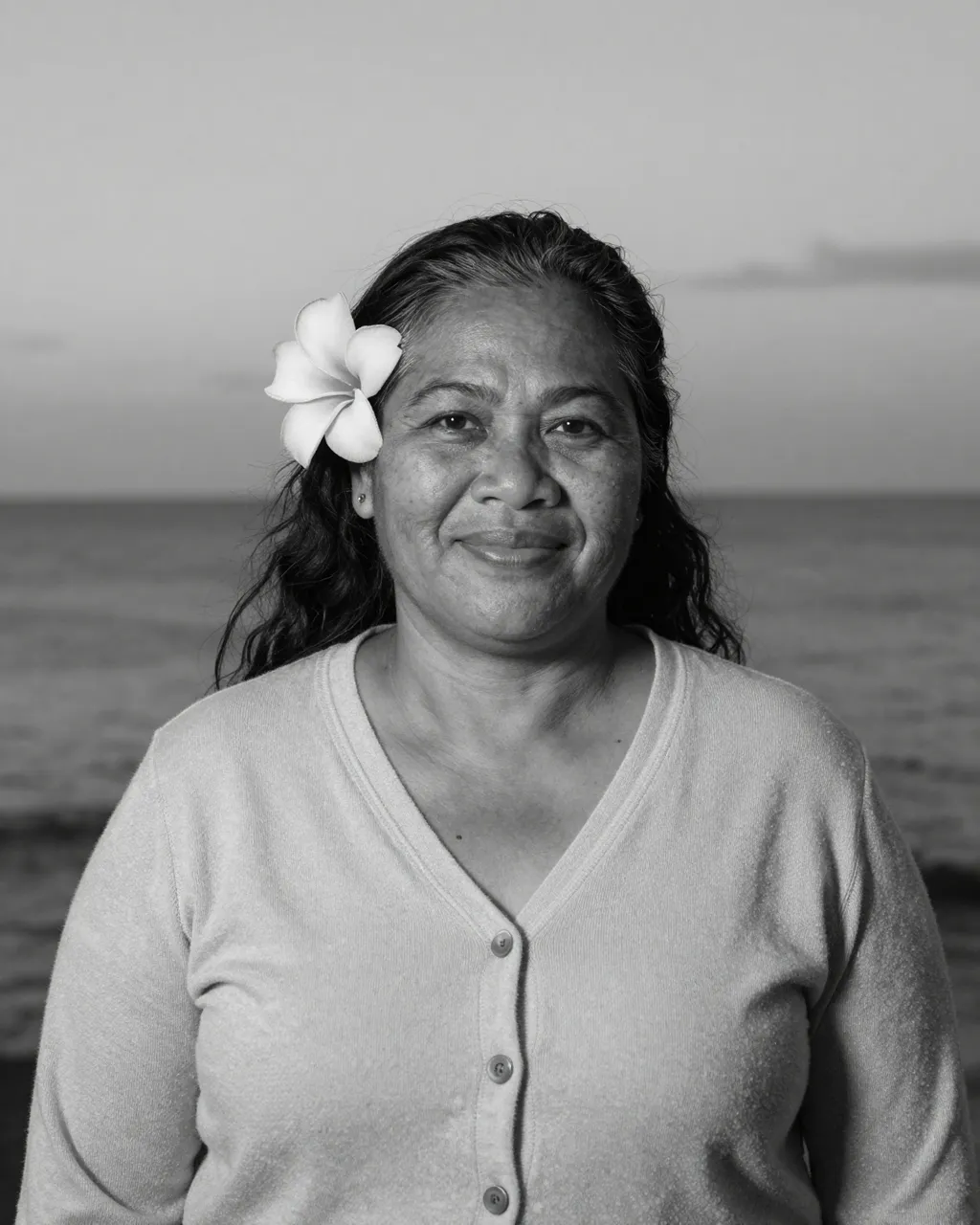 Black and white portrait of a smiling older woman with a plumeria flower behind her ear standing by the sea at dusk