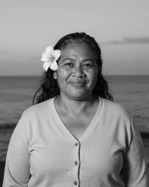 Black and white portrait of a smiling older woman with a plumeria flower behind her ear standing by the sea at dusk