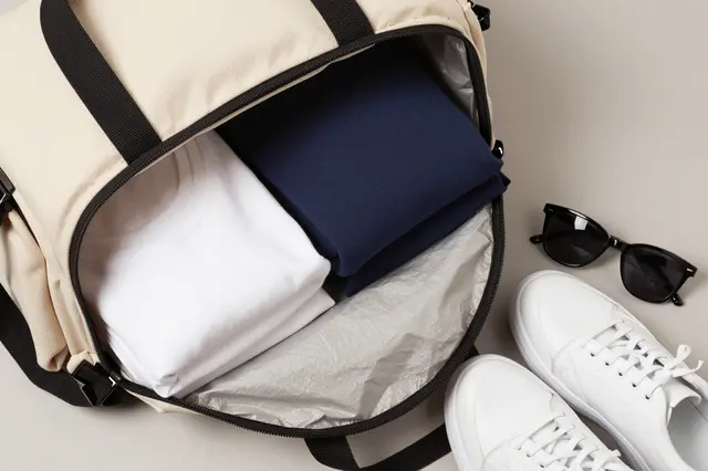 Top down view of an open canvas duffel with packed clothes, white sneakers and sunglasses on a neutral gray surface