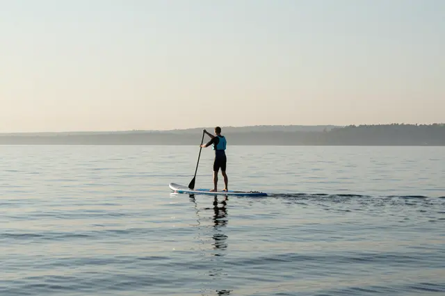A stand up paddleboarder glides across calm lake water at hazy sunrise, viewed from a distance.