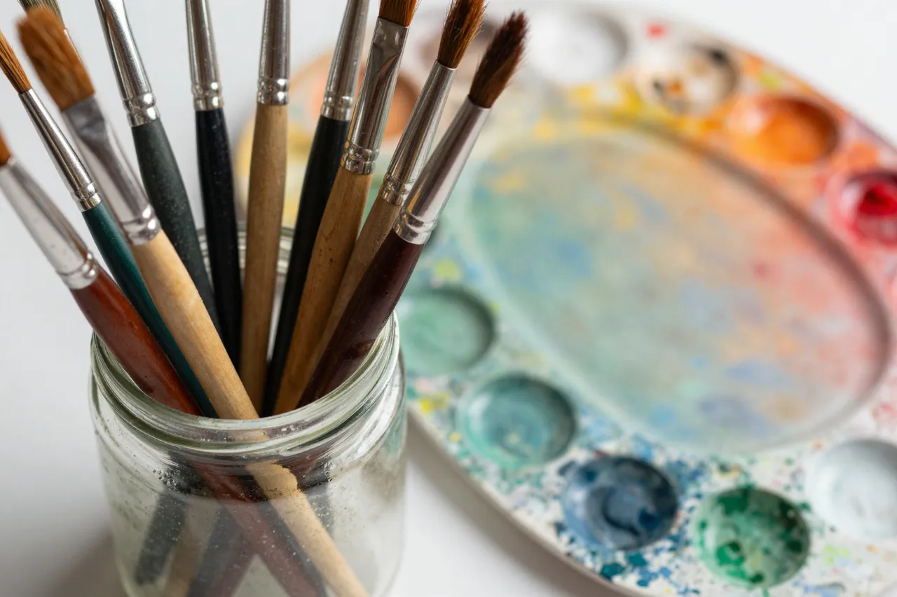 A glass jar holding assorted paintbrushes sits next to a used multicolor paint palette on a white surface