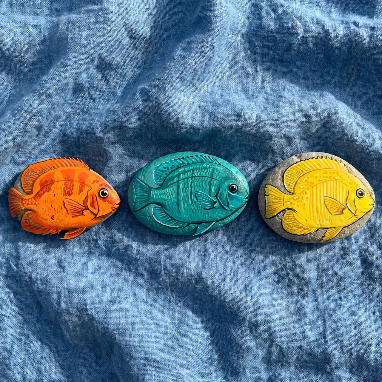 Three river stones painted as orange, teal and yellow tropical fish rest on crumpled blue linen