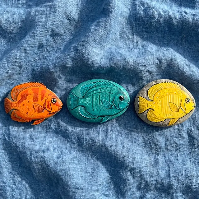 Three river stones painted as orange, teal and yellow tropical fish rest on crumpled blue linen