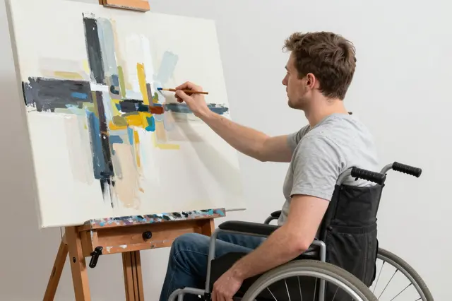 Side view of a male painter in a wheelchair adding details to an abstract canvas on an easel