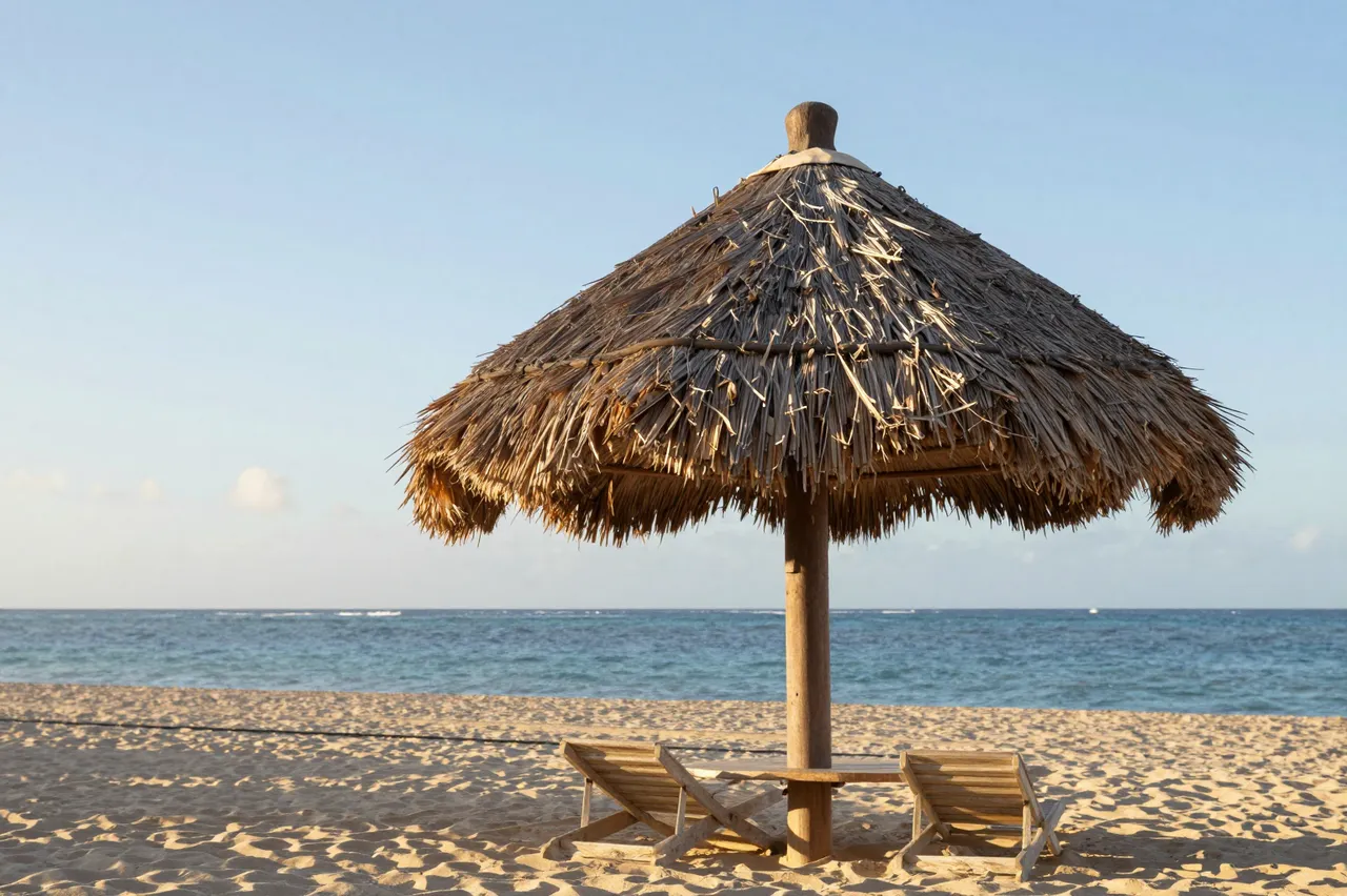 A palm thatched palapa sits over beach lounge chairs on a sandy Caribbean beach at sunrise beside calm ocean