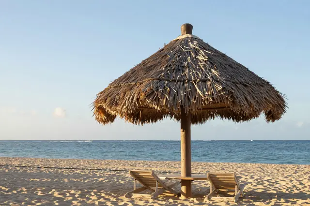 A palm thatched palapa sits over beach lounge chairs on a sandy Caribbean beach at sunrise beside calm ocean