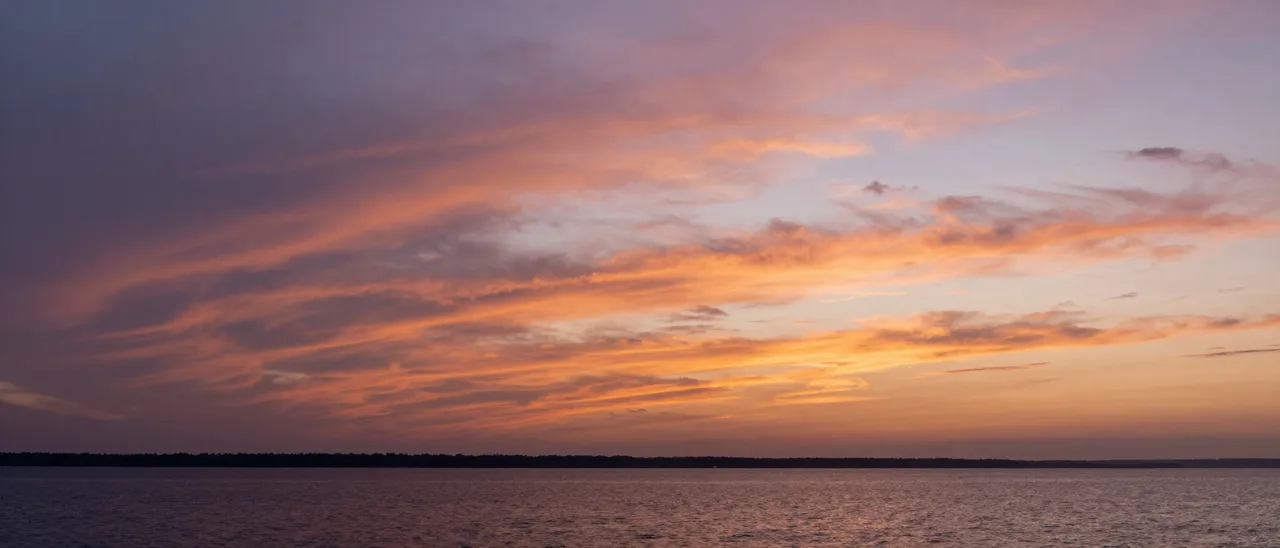 Layered orange and violet sunset clouds stretch over calm bay water and a distant tree-lined shore